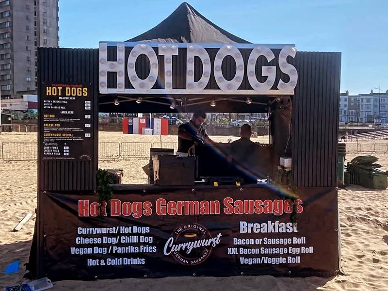 The Currywurst pop-up stall with HOT DOGS light-up sign on a beach