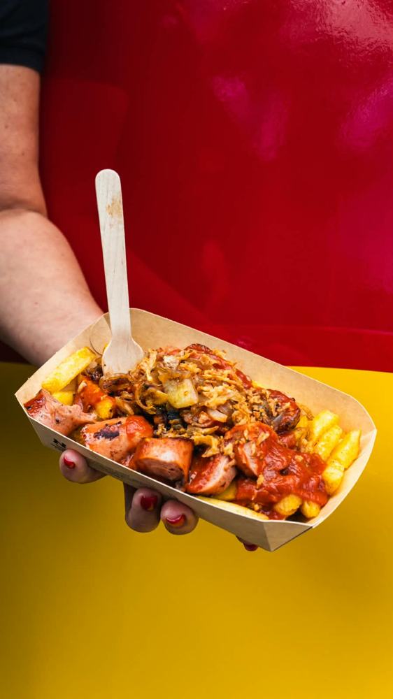 A tray of authentic Berlin-style currywurst and fries held against red and yellow